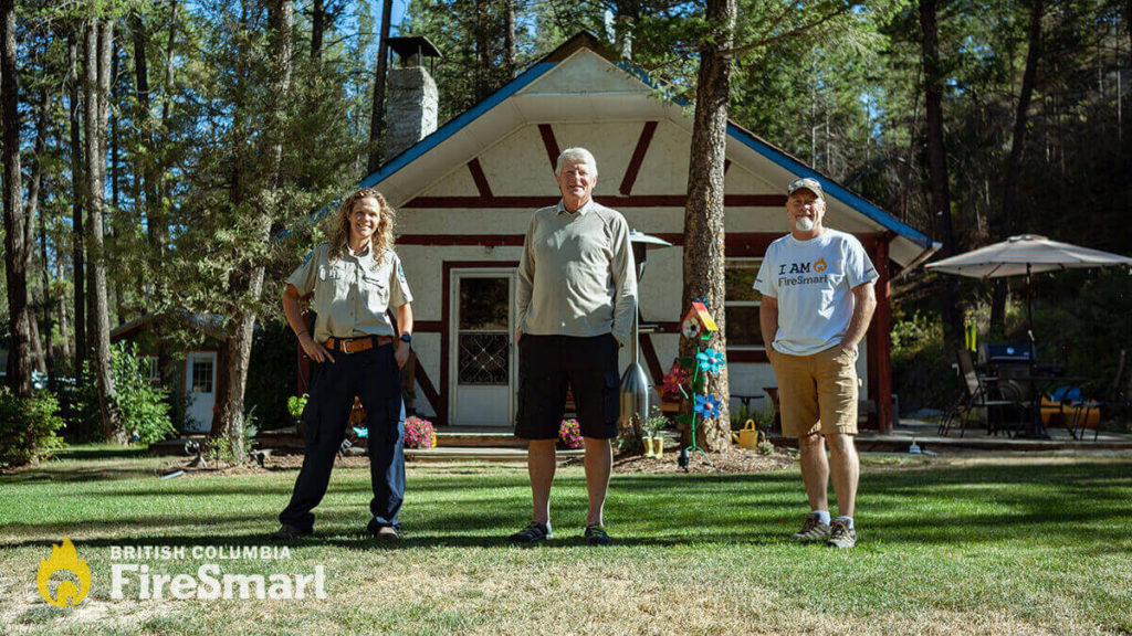 Three people stand outside a FireSmart-ed house in B.C. There are trees around the house. One person is wearing a uniform, the other appears to be the homeowner, and the third is wearing an "I am FireSmart" t-shirt.
