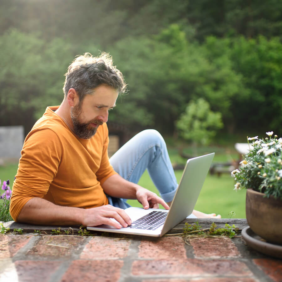 A man sitting barefoot outside, presumably in his backyard. He is looking intently at a laptop, and the sun is shining from behind him.