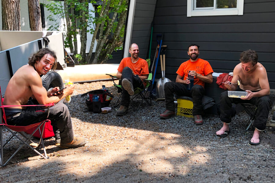 The Garibaldi Tree crew taking a break from tree work in Whistler. They're sitting in a client's driveway. Everyone is smiling and laughing, and it's a sunny day.