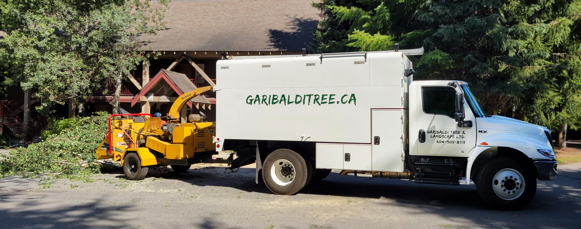 The Garibaldi Tree work truck outside a building in Whistler, B.C. The truck is a white dump truck with "garibalditree.ca" across the side in green lettering. There is a yellow chipper attached to the truck, with some branches sticking out the back of it.
